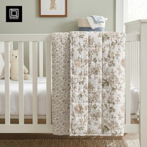 Könnte beinhalten: Ein weißes Babybett mit einer floral gemusterten Steppdecke, die über die Seite drapiert ist. Die Steppdecke hat ein sich wiederholendes Muster aus beigen und grünen Blumen auf weißem Grund. Ein Teddybär sitzt im Bett.