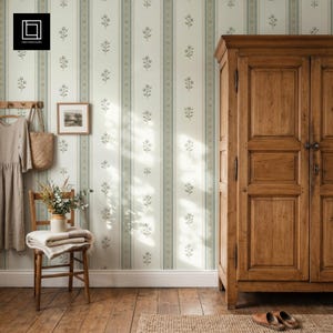 Op de afbeelding: Een hoge houten kledingkast met dubbele deuren staat naast een houten stoel met een opgevouwen deken en een vaas met droogbloemen. Een beige jurk hangt aan een haak en een ingelijste afbeelding hangt aan behang met groene bloemenstrepen.