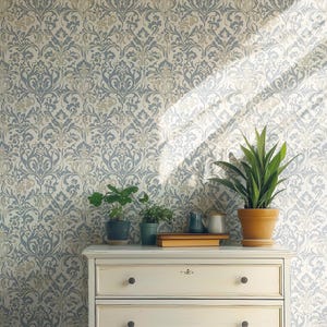 May include: A cream-colored dresser with two drawers, topped with potted plants and books, sits against a wallpaper backdrop. The wallpaper features a repeating floral pattern in shades of blue, gray, and cream.