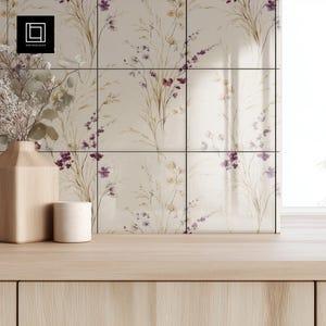 May include: A light wood vase and a small white ceramic cup sit on a light wood surface. Behind them is a wall of square tiles with a floral pattern in beige, purple, and brown tones. The tiles have black lines separating them.