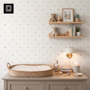 May include: A nursery room featuring a light-coloured wall with a repeating pattern of small pink hearts. Two wooden shelves hold two teddy bears, a framed illustration, books, and a small potted plant. A woven changing basket sits on a dresser.