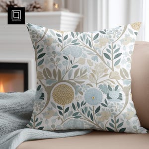 May include: A white throw pillow with a floral pattern featuring blue, green, and gold flowers and leaves. The pillow is sitting on a couch with a light blue throw blanket.