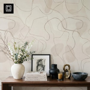 May include: A wooden table displays home decor items against a neutral-toned abstract wallpaper. Items include a white vase with flowers, framed artwork, books, a black vase, a candle, a gold chain sculpture, and a grey bowl.