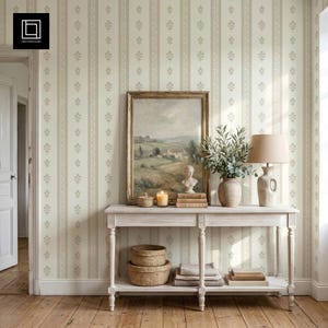 May include: A light-colored wooden console table with a lower shelf, holding a framed landscape painting, a ceramic vase with greenery, a lamp, and two woven baskets. The wall features vertical striped wallpaper with small floral motifs.