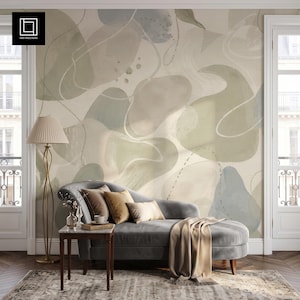 Könnte beinhalten: Eine graue Samt-Chaiselongue mit einer beigen Decke, zwei braunen Kissen und einem kleinen Beistelltisch. Die Wand zeigt eine großflächige Tapete mit abstrakten organischen Formen in gedämpften Salbeigrün-, Grau- und Cremetönen mit Linien.