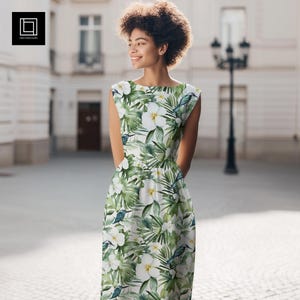 May include: Sleeveless white dress with a tropical floral print featuring white flowers, green leaves, and blue birds. The dress has a scoop neckline and a fitted waist. The model is standing outside in front of a building.