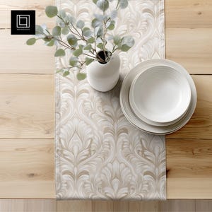 May include: A beige and white patterned table runner with a vase of greenery and white plates. The runner features a swirling, floral design. The plates are stacked on top of each other. The vase is white and has a unique shape.
