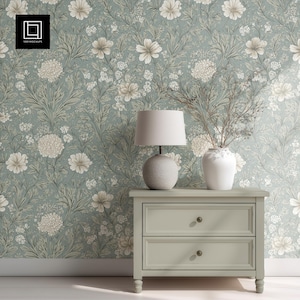 May include: A light green nightstand with two drawers, a round lamp, and a white vase with branches. The background features floral wallpaper in shades of green, white, and beige. The nightstand has two drawers with round knobs.