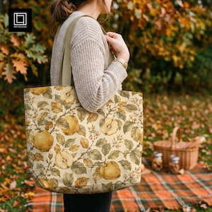 May include: A large tote bag featuring a repeating botanical print of yellow apples, green leaves, and small white blossoms on a cream background. The bag has wide, sturdy beige fabric straps and a rectangular shape with a flat bottom base.