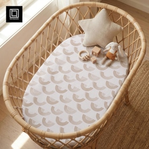 May include: A woven rattan bassinet with a patterned sheet, star-shaped pillow, and stuffed bunny. The sheet features a repeating design of beige semi-circles on a white background. Wooden toys are also present.