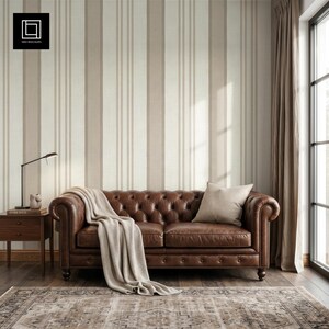 Könnte beinhalten: Ein braunes, getuftetes Ledersofa mit gerollten Armlehnen und Knopfdetails, drapiert mit einer hellbeigen Decke. Ein einzelnes beiges Kissen liegt auf dem Sofa. Die Wand zeigt vertikale Streifen in abwechselnden Creme- und Hellbrauntönen.