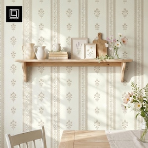 May include: A light-colored wooden wall shelf holds two white ceramic pitchers, a stack of three books, two framed botanical prints, and a wooden cutting board. The shelf is mounted against a cream-colored wallpaper with a repeating floral pattern.