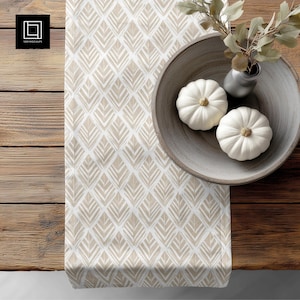 May include: A beige and white patterned table runner with a diamond leaf design. Two white pumpkins sit in a wooden bowl with a small vase of greenery. The table is made of wood.