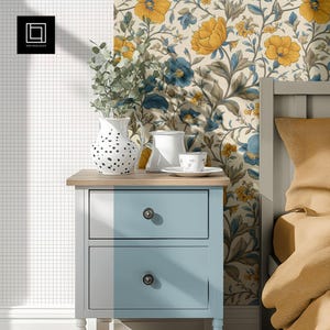May include: A blue nightstand with two drawers and black knobs. The nightstand has a wood top with a white vase with black polka dots, a white pitcher, and a white teacup and saucer on it. The nightstand is in front of a wall with a floral wallpaper with yellow and blue flowers.