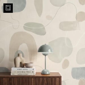 May include: A gray desk lamp with a rounded shade sits on a dark wood surface. Beside it are books, a beige vase, and a small bowl. The background features a cream-colored wall with abstract shapes in muted tones of gray and beige.
