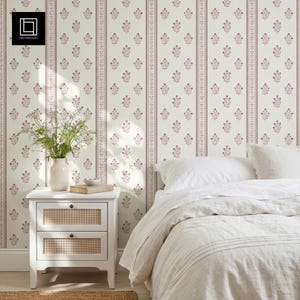 Può includere: Carta da parati con un motivo ripetitivo di piccoli fiori rosa disposti in colonne verticali separate da sottili linee a righe su uno sfondo color crema. Il design è mostrato su una parete dietro un comodino bianco e un letto.