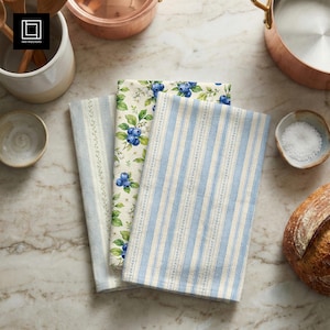 May include: Three rectangular kitchen towels featuring varied patterns. One towel displays a light blue and white vertical stripe design, another shows a repeating blueberry and green leaf print, and the third features a subtle light blue pinstripe.