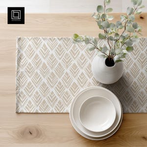 Puede incluir: Una mesa con un camino de mesa beige y blanco estampado, platos de cerámica blancos y un jarrón blanco con follaje verde. La mesa es de madera clara.