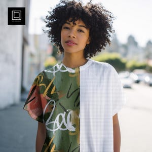 May include: A short-sleeved shirt featuring a split design. The left side displays an olive green background with abstract white, yellow, and orange brushstroke patterns. The right side features a clean, white fabric with a fine black grid pattern.