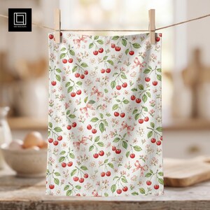 Puede incluir: Un paño de cocina blanco con un patrón repetitivo de cerezas rojas sobre tallos verdes, pequeñas flores de cerezo blancas y lazos decorativos de color rosa claro. La tela es rectangular y cuelga de una pinza de madera.