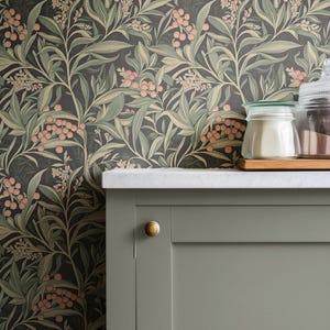May include: A close-up of a kitchen scene featuring a sage green cabinet with a gold knob, a white countertop, and a floral wallpaper background. The wallpaper has a dark background with green leaves and pink berries. Glass jars are on the countertop.