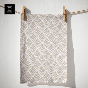 May include: A white and beige patterned kitchen towel hanging from a clothesline. The towel features a repeating diamond pattern with leaf-like shapes in beige. The towel is held by two wooden clothespins against a white wall.