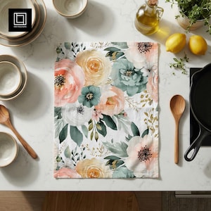 May include: A white floral kitchen towel with a watercolor design featuring peach, yellow, and teal flowers and green leaves. The towel is rectangular and lies flat on a white surface, surrounded by kitchen items.