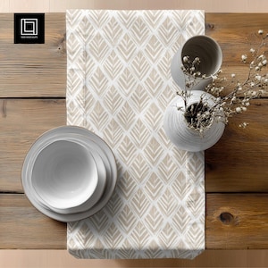 May include: A table setting featuring a cream-colored table runner with a geometric leaf pattern. The runner is set on a wooden table with white ceramic plates, bowls, and vases. A sprig of white flowers adds a touch of elegance.