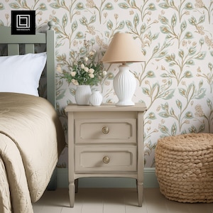 May include: A bedroom scene featuring a light gray nightstand with two drawers, topped with a white lamp and vases. The bed has a beige comforter, and the wallpaper has a floral pattern in green and gold. A woven pouf sits on the floor.