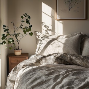 May include: A bedroom interior with a bed dressed in a floral duvet and pillowcases. A small wooden bedside table supports a potted plant. Sunlight streams through a window, creating shadows on the bedding and wall.