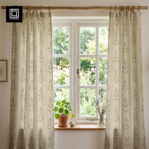 Op de afbeelding: Beige linnen gordijnen met een herhalend patroon van bruine bladeren en bessen. De gordijnen hangen aan houten ringen aan een houten roede en omlijsten een raam met uitzicht op een tuin. Een potplant en een vaas met bloemen staan op de vensterbank.
