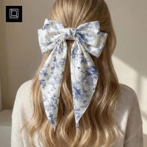 May include: A large white hair bow with a blue floral pattern is secured in the hair. The bow is made of a silky material and has long, flowing tails. The floral design features blue flowers and leaves on a white background. The hair accessory adds a touch of elegance.