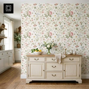 May include: A cream sideboard with a light wood top and brass handles. The wall behind is covered in floral wallpaper with pink roses and green leaves. A vase of white flowers and a bowl of lemons sit on top of the sideboard.