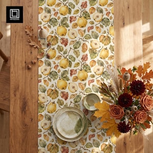 May include: A table runner featuring a repeating pattern of yellow quince fruits and green leaves on a cream background. The runner is decorated with a stack of two speckled ceramic plates and a floral arrangement of roses and dried foliage.