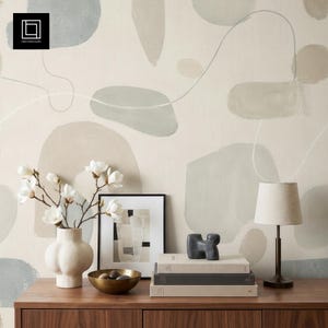 May include: A wooden cabinet displays a white vase with white flowers, a framed abstract art print, a brass bowl with dark spheres, and a stack of books. A small lamp and a dark sculpture sit on top. The background features a beige wall with abstract shapes.