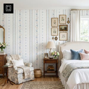 Puede incluir: Un dormitorio con papel tapiz de rayas verticales azules y beige, un sillón estampado con un cojín blanco, una mesita de noche de madera con lámpara y libros, y una cama con ropa de cama blanca, cojines azules y rosas.