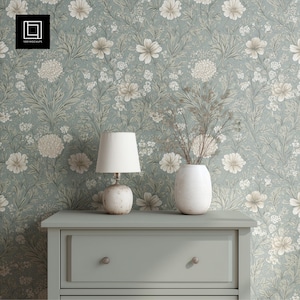 May include: A light blue dresser with a white lamp and a white vase with dried plants. The background is a floral wallpaper in shades of blue, white, and gray. The lamp has a white lampshade and a ceramic base.