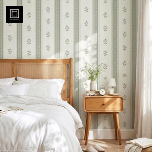 Può includere: Una camera da letto con un letto dotato di testiera in canna di legno chiaro, biancheria bianca e un comodino in legno con lampada e orologio. La parete è rivestita con carta da parati a righe verticali e un piccolo motivo floreale verde.