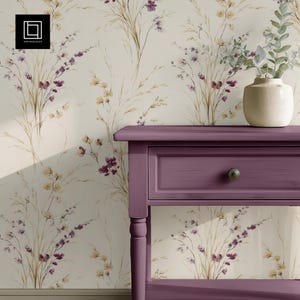 May include: A purple side table with a drawer and a cream-colored vase holding greenery. The table is set against a floral wallpaper with beige, purple, and yellow flowers. The wallpaper has a repeating pattern of wildflowers.