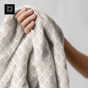 Puede incluir: Una tela beige y blanca estampada es sostenida en una mano. La tela presenta un diseño geométrico repetido de formas de hojas. La tela es suave y parece estar drapeada. El logotipo "1001 Mockups" está en la esquina superior izquierda.