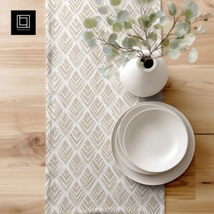 Op de afbeelding: Een tafelsetting met een beige en wit patroon tafelloper, witte borden, een witte vaas en groen. De tafelloper heeft een geometrisch bladontwerp. De borden zijn opgestapeld en de vaas is rond. De setting staat op een houten tafel.