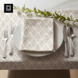 May include: A table setting featuring a white plate with a folded napkin, a knife, a spoon, and two forks. The napkin and table runner have a repeating geometric pattern in beige and white. A sprig of greenery adds a natural touch.