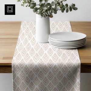 May include: A beige and white patterned table runner with a geometric design. A stack of white plates and a white vase with greenery are on the wooden table. The table runner has a repeating pattern of stylized leaf shapes.