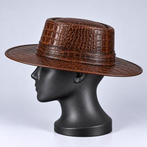 Op de afbeelding: Een bruine fedora-hoed met een gestructureerd krokodillenleerpatroon over de kroon en de brede rand. De hoed heeft een gestructureerd ontwerp met een bijpassende bruine band rond de basis van de kroon, met een consistente schubbenafwerking.