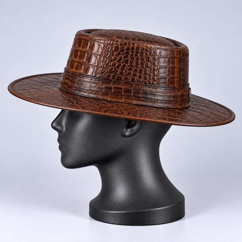Op de afbeelding: Een bruine fedora-hoed met een gestructureerd krokodillenleerpatroon over de kroon en de brede rand. De hoed heeft een gestructureerd ontwerp met een bijpassende bruine band rond de basis van de kroon, met een consistente schubbenafwerking.