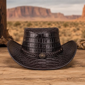 May include: A dark brown cowboy hat featuring a textured crocodile skin pattern across the crown and brim. The hat is accented with a braided leather band and a central metallic circular emblem displaying a five-pointed star design.