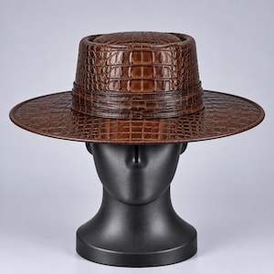Op de afbeelding: Een bruine hoed met een gestructureerd krokodillenleerpatroon, voorzien van een platte kroon en een brede, horizontale rand. De hoed heeft een bijpassende bruine band rond de basis van de kroon, wat zorgt voor een uniforme uitstraling over het geheel.