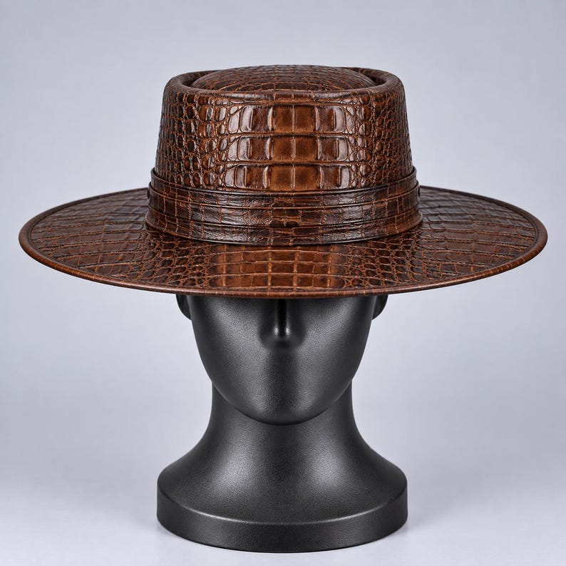 Op de afbeelding: Een bruine hoed met een gestructureerd krokodillenleerpatroon, voorzien van een platte kroon en een brede, horizontale rand. De hoed heeft een bijpassende bruine band rond de basis van de kroon, wat zorgt voor een uniforme uitstraling over het geheel.