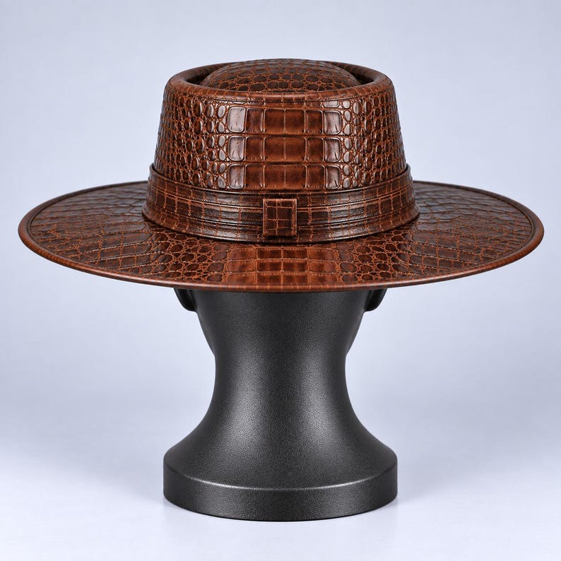 Op de afbeelding: Een bruine fedora hoed met een textuur van krokodillenleer over het gehele oppervlak. De hoed heeft een platte bovenkant, een brede rand en een bijpassende bruine band met een klein rechthoekig detail in het midden aan de basis van de kroon.