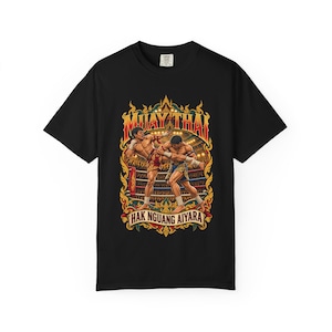 May include: A black t-shirt featuring a graphic print of two Muay Thai fighters in a ring. The design includes the text "MUAY THAI" at the top and "HAK NGUANG AIYARA" at the bottom, surrounded by ornate gold and red decorative borders.